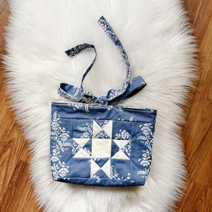 May include: A quilted fabric tote bag featuring a blue and white floral damask pattern with a central white star block design. The bag includes two matching fabric handles tied in a knot at the top, showcasing a structured, stitched construction.