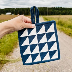 May include: A square, quilted potholder with a navy blue border and handle. The potholder features a pattern of white and navy blue triangles. The potholder is held up against a backdrop of a dirt road and green fields.