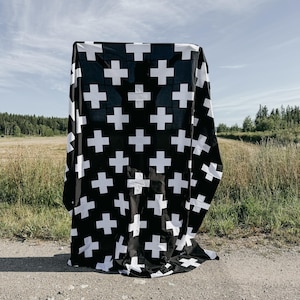May include: A black and white patterned textile with a repeating cross design. The fabric is draped over an unseen object, creating a visually striking contrast against a natural outdoor backdrop of fields and sky.