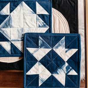 以下が含まれることがあります： インディゴブルーと白の幾何学的な星柄の正方形のキルトコースター。コースターは濃い青色の縁取りがあり、木製の表面に配置されています。デザインは伝統的なキルトを彷彿とさせます。