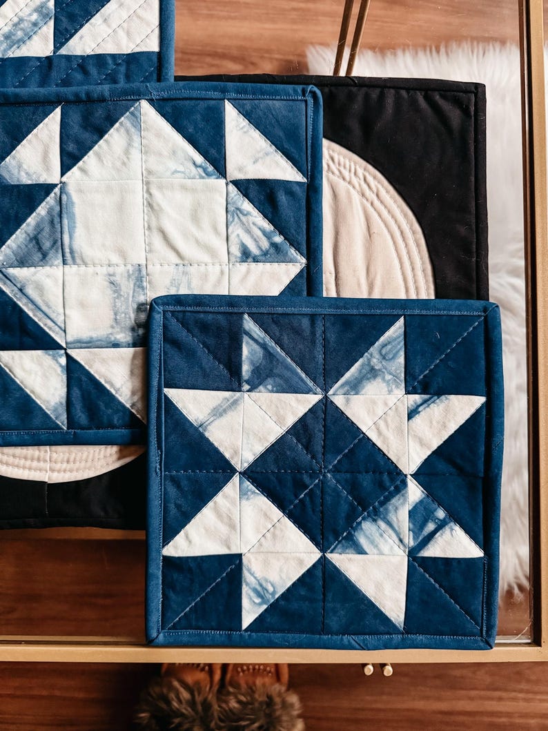 以下が含まれることがあります： インディゴブルーと白の幾何学的な星柄の正方形のキルトコースター。コースターは濃い青色の縁取りがあり、木製の表面に配置されています。デザインは伝統的なキルトを彷彿とさせます。