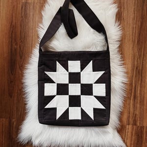 May include: Black and white quilted shoulder bag with a starburst and checkerboard pattern. The bag has a long, black strap and is displayed on a white, fluffy rug. The bag is a square shape.