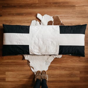 May include: A long, rectangular body pillow with a black and white design. The pillow has a black border with a white cross in the center. The pillow is resting on a cowhide rug on a wooden floor.