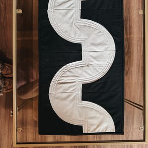 May include: Black and white quilted table runner with a unique, curved design. The runner features a bold, contrasting pattern of a white, wave-like shape against a black background. The design is centered on a glass table with gold trim.