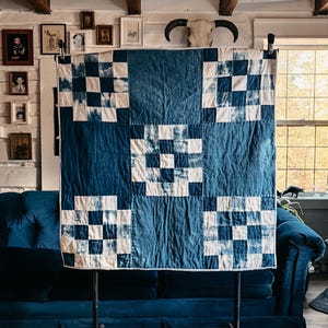 May include: A blue and white quilt with a checkerboard pattern is displayed on a black metal stand. The quilt is made of fabric with a dark blue background and white squares. The room has a rustic aesthetic with a blue velvet sofa and wooden ceiling beams.
