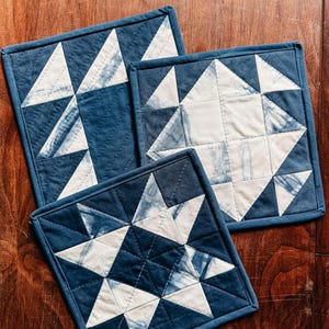 以下が含まれることがあります： 星型のデザインが施された、正方形のキルト鍋つかみ3つ。鍋つかみは藍色で、白い星模様が施されています。縁は、お揃いの青い縁取りでトリミングされています。鍋つかみは木製の表面に配置されています。