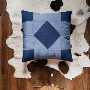 May include: A square, patchwork pillow in shades of blue, featuring a central diamond design. The pillow is placed on a cowhide rug with brown and black markings, creating a rustic aesthetic. The pillow's design is geometric and modern.
