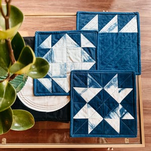 Pode incluir: Um conjunto de panos de prato quadrados acolchoados, feitos à mão, em tons de azul índigo e branco. Apresentam padrões geométricos de estrelas e triângulos. Os panos de prato estão dispostos sobre uma mesa com tampo de vidro.