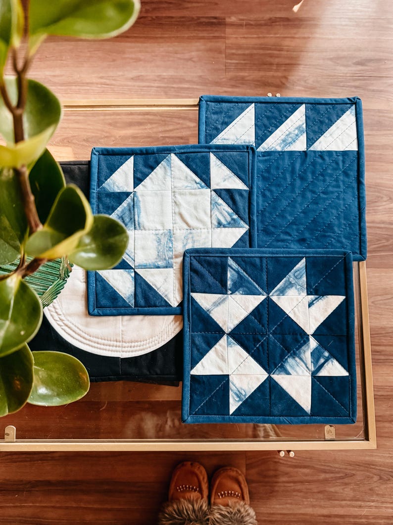 以下が含まれることがあります： インディゴブルーと白の正方形のキルト鍋つかみのコレクション。鍋つかみは、幾何学的な星と三角形のパターンが特徴です。手作りのアイテムは、ガラス張りのテーブルに配置されています。