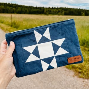 Puede incluir: Bolsa acolchada azul marino con un dise&ntilde;o de estrella blanca. Tiene cierre de cremallera y una peque&ntilde;a etiqueta de cuero marr&oacute;n con la inscripci&oacute;n "EXSHAW". El fondo muestra un campo y &aacute;rboles borrosos.