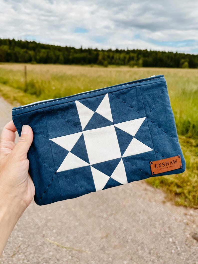Puede incluir: Bolsa acolchada azul marino con un dise&ntilde;o de estrella blanca. Tiene cierre de cremallera y una peque&ntilde;a etiqueta de cuero marr&oacute;n con la inscripci&oacute;n "EXSHAW". El fondo muestra un campo y &aacute;rboles borrosos.