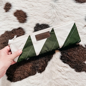 May include: A rectangular pencil case with a white and green chevron pattern. The case has a zip closure and a small brown leather patch with text. The case is held by a hand.