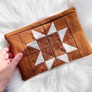 May include: A brown and white quilted clutch with a star pattern. The rectangular bag features a geometric design with brown squares and white triangles. The clutch is held by a hand and rests on a white, fluffy surface.