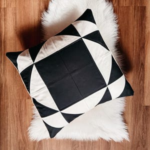 Op de afbeelding: Vierkant decoratief kussen met een geometrisch zwart-wit ontwerp. Het kussen ligt op een witte imitatiebontvloerkleed, tegen een houten vloerachtergrond. Het patroon heeft driehoeken en vierkanten.