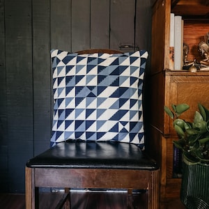Puede incluir: Un cojín decorativo cuadrado con un patrón geométrico de triángulos blancos y azules. Está sobre una silla de madera con asiento de cuero negro. El fondo incluye una pared de madera oscura y una estantería de madera.