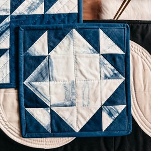 以下が含まれることがあります： 幾何学模様の正方形のキルト鍋つかみ2つ。デザインは白とインディゴブルーの三角形と正方形が特徴です。鍋つかみは濃い青色の縁取りがあります。鍋つかみは黒い表面にあります。