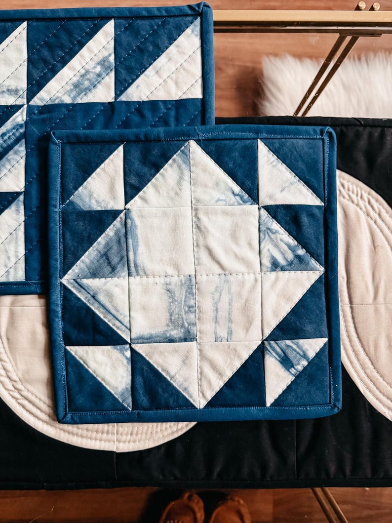 以下が含まれることがあります： 幾何学模様の正方形のキルト鍋つかみ2つ。デザインは白とインディゴブルーの三角形と正方形が特徴です。鍋つかみは濃い青色の縁取りがあります。鍋つかみは黒い表面にあります。