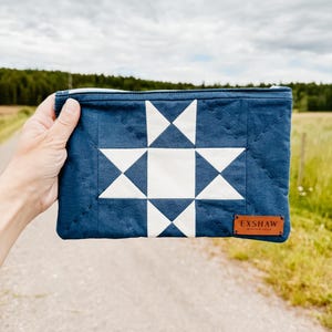 Puede incluir: Un estuche acolchado azul marino con un dise&ntilde;o de estrella blanca. El estuche rectangular tiene cierre de cremallera y una peque&ntilde;a etiqueta de cuero marr&oacute;n con la inscripci&oacute;n "EXSHAW". El fondo es una escena borrosa al aire libre.