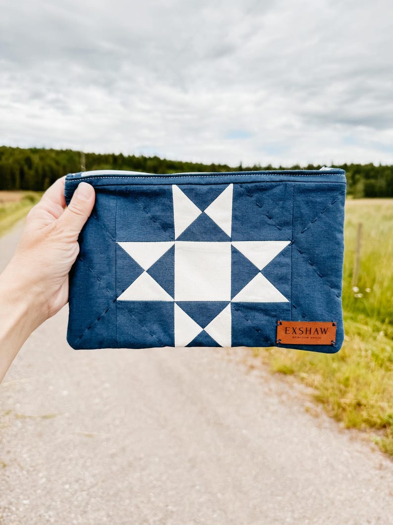 Puede incluir: Un estuche acolchado azul marino con un dise&ntilde;o de estrella blanca. El estuche rectangular tiene cierre de cremallera y una peque&ntilde;a etiqueta de cuero marr&oacute;n con la inscripci&oacute;n "EXSHAW". El fondo es una escena borrosa al aire libre.