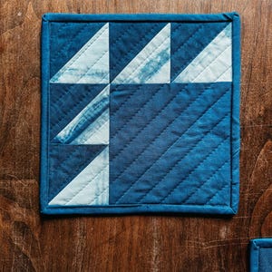 以下が含まれることがあります： 幾何学模様のキルティングコースター。コースターは濃い青色の縁取りと、青と白の三角形のパターンが特徴です。生地は質感があり、木製の表面に置かれています。