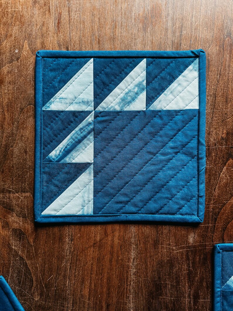 以下が含まれることがあります： 幾何学模様のキルティングコースター。コースターは濃い青色の縁取りと、青と白の三角形のパターンが特徴です。生地は質感があり、木製の表面に置かれています。