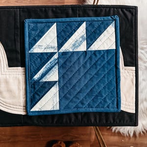 以下が含まれることがあります： 青と白の色合いの正方形のキルト生地で、幾何学模様が特徴です。 黒い表面にクリーム色の縁取りが施されています。 コースターや小さなマットなど、装飾品である可能性があります。