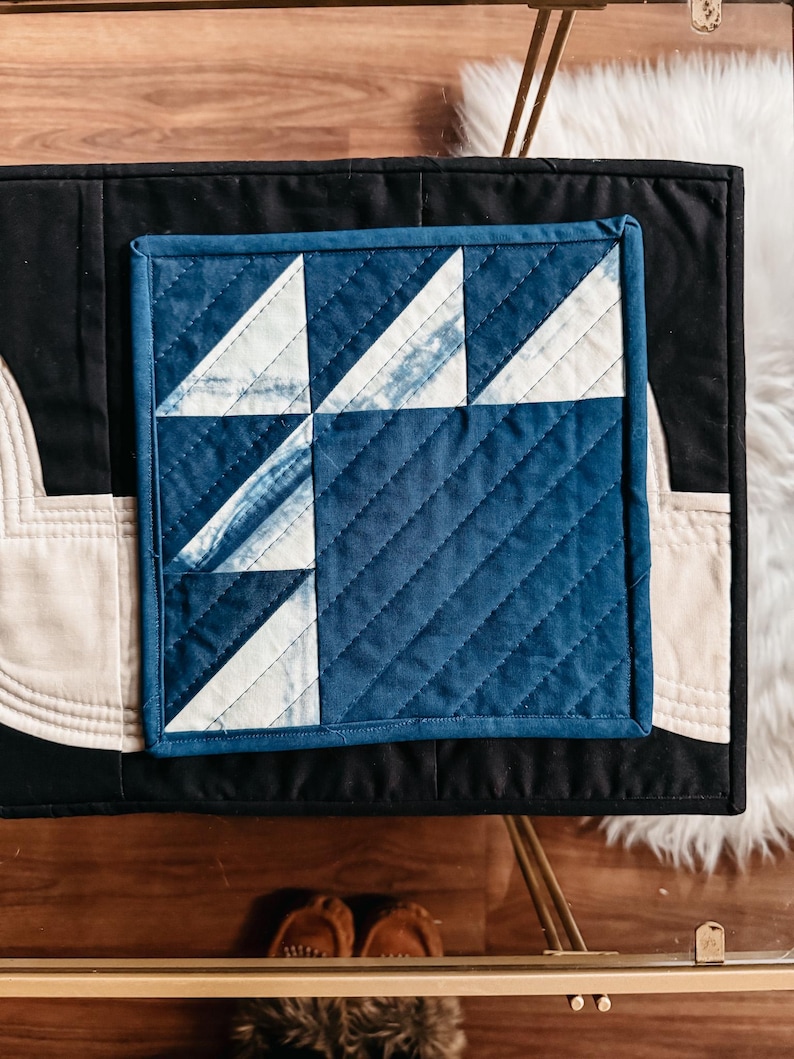 以下が含まれることがあります： 青と白の色合いの正方形のキルト生地で、幾何学模様が特徴です。 黒い表面にクリーム色の縁取りが施されています。 コースターや小さなマットなど、装飾品である可能性があります。