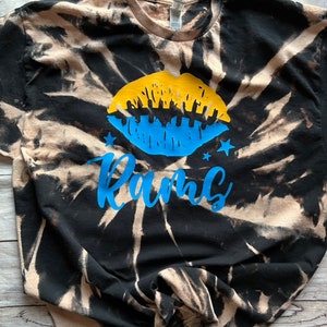 May include: Black and white tie-dye t-shirt with a blue and yellow lip design and the word "Rams" in blue lettering.