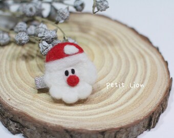Puede incluir: Una pinza para el pelo de fieltro de Papá Noel, con gorro rojo, barba blanca y nariz roja. La pinza es plateada y brillante. La pinza está sobre una rodaja de madera con el texto "Petit Lion".