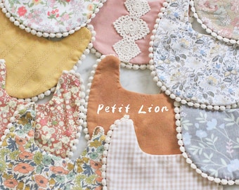 Può includere: Una collezione di bavaglini per bambini in vari tessuti floreali e fantasia, tra cui rosa, giallo e marrone. Ogni bavaglino è rifinito con pompon bianchi e alcuni presentano dettagli in pizzo o a quadretti. Il testo "Petit Lion" è visibile.