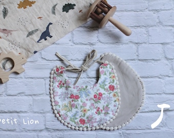 Può includere: Due bavaglini per bambini, uno floreale e uno beige tinta unita, con bordatura a pompon color crema. Sono visibili anche un sonaglio in legno e un tessuto a tema dinosauri. Il testo "Petit Lion" è stampato in basso a sinistra.