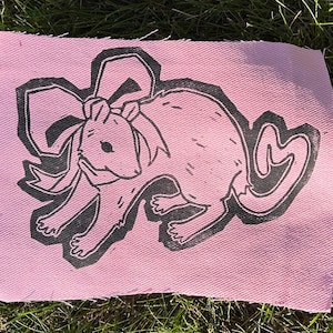 Peut inclure: Un écusson en tissu rose rectangulaire présentant une impression en bloc d'encre noire représentant une souris portant un grand nœud sur la tête. La souris est représentée de profil avec une longue queue courbée et des lignes de fourrure texturées.