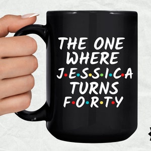 Op de afbeelding: Zwarte koffiemok met witte tekst die "The One Where Jessica Turns Forty" luidt, met gekleurde stippen die de klinkers in de woorden vervangen. De mok staat op een witte achtergrond.