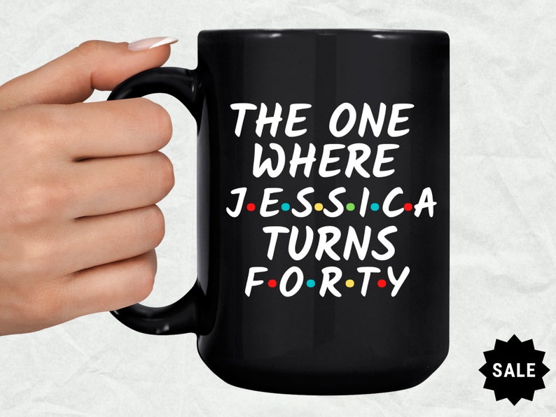 Op de afbeelding: Zwarte koffiemok met witte tekst die "The One Where Jessica Turns Forty" luidt, met gekleurde stippen die de klinkers in de woorden vervangen. De mok staat op een witte achtergrond.