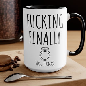 May include: White ceramic coffee mug with a black rim and handle. The mug has black text that reads "FUCKING FINALLY" with a diamond ring illustration and the text "MRS. THOMAS" below.