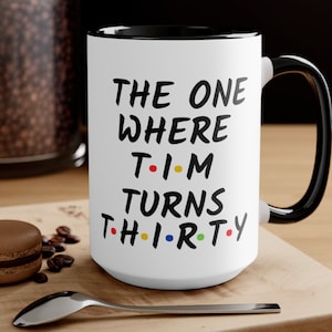 May include: White ceramic mug with a black rim and handle. The mug has black text that reads "THE ONE WHERE T-I-M TURNS TH-I-R-TY". The letters "T-I-M" are in red, yellow, and blue, and the letters "TH-I-R-TY" are in red, yellow, blue, and green.