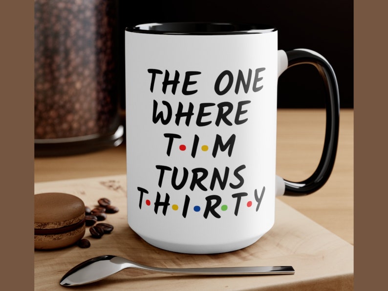 May include: White ceramic mug with a black rim and handle. The mug has black text that reads "THE ONE WHERE T-I-M TURNS TH-I-R-TY". The letters "T-I-M" are in red, yellow, and blue, and the letters "TH-I-R-TY" are in red, yellow, blue, and green.