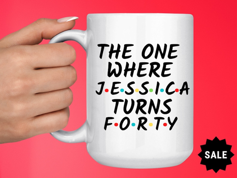 Op de afbeelding: Witte keramische mok met zwarte tekst die "The One Where Jessica Turns Forty" luidt, met gekleurde stippen die de klinkers vervangen. De mok wordt vastgehouden door een hand.