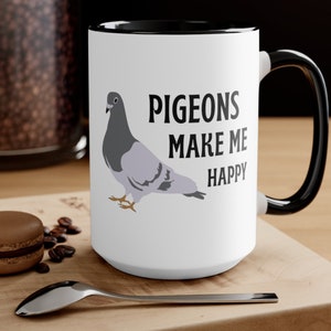 May include: White ceramic mug with a black interior and handle. The mug features a gray pigeon illustration and the text "PIGEONS MAKE ME HAPPY" in black. The mug is on a wooden surface with a spoon and a pastry.