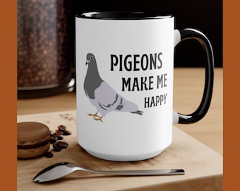 May include: White ceramic mug with a black interior and handle. The mug features a gray pigeon illustration and the text "PIGEONS MAKE ME HAPPY" in black. The mug is on a wooden surface with a spoon and a pastry.