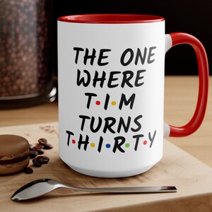 May include: White ceramic coffee mug with a red rim and handle. The mug has black text that reads "THE ONE WHERE T-I-M TURNS TH-I-R-T-Y". The "I"s are replaced with colorful dots.
