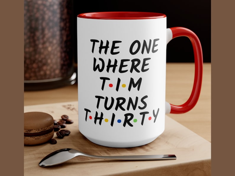 May include: White ceramic coffee mug with a red rim and handle. The mug has black text that reads "THE ONE WHERE T-I-M TURNS TH-I-R-T-Y". The "I"s are replaced with colorful dots.