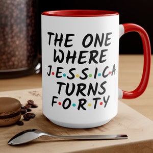 Op de afbeelding: Witte keramische mok met een rode rand en handvat. De mok heeft zwarte tekst die "The One Where Jessica Turns Forty" luidt, met kleurrijke stippen die de klinkers in de naam en leeftijd vervangen.
