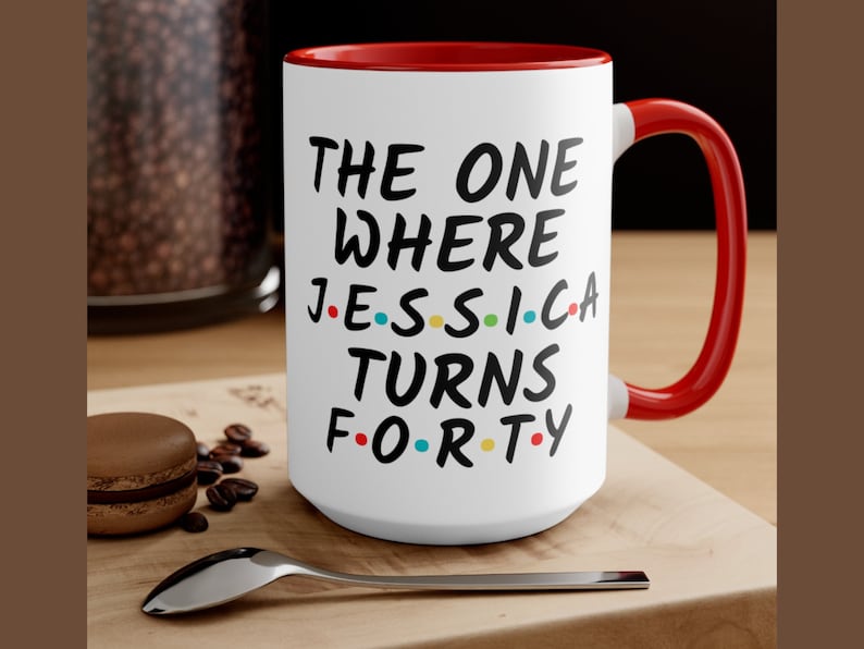 Op de afbeelding: Witte keramische mok met een rode rand en handvat. De mok heeft zwarte tekst die "The One Where Jessica Turns Forty" luidt, met kleurrijke stippen die de klinkers in de naam en leeftijd vervangen.