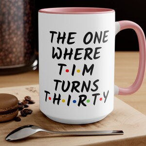 May include: White ceramic coffee mug with a pink rim and handle. The mug has black text that reads "THE ONE WHERE T-I-M TURNS TH-I-R-TY".