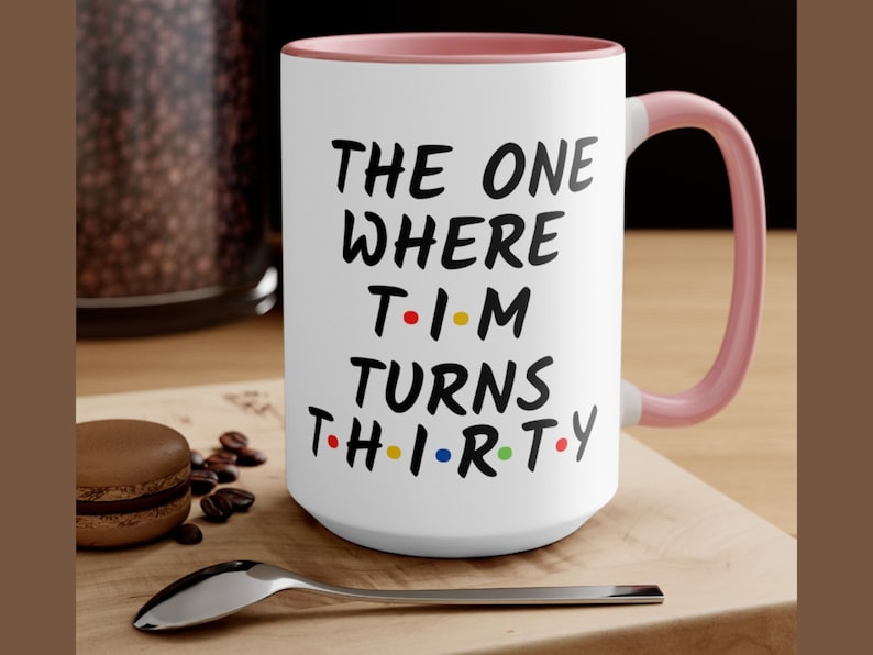 May include: White ceramic coffee mug with a pink rim and handle. The mug has black text that reads "THE ONE WHERE T-I-M TURNS TH-I-R-TY".