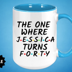 Op de afbeelding: Witte keramische mok met een blauwe rand en handvat. De mok heeft zwarte tekst die "The One Where Jessica Turns Forty" luidt, met gekleurde stippen die de klinkers vervangen.