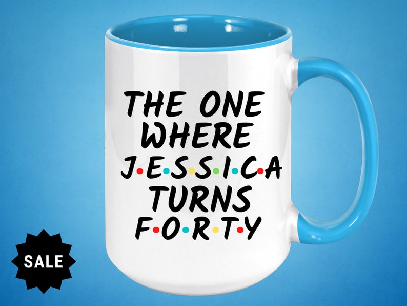 Op de afbeelding: Witte keramische mok met een blauwe rand en handvat. De mok heeft zwarte tekst die "The One Where Jessica Turns Forty" luidt, met gekleurde stippen die de klinkers vervangen.