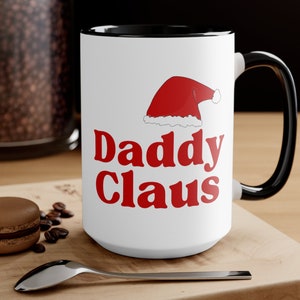 May include: White ceramic mug with a black rim and handle. The mug has a red and white Santa hat graphic and the text "Daddy Claus" in red letters.