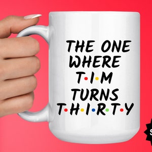 May include: White ceramic mug with black text that reads "The One Where Tim Turns Thirty" with colorful dots replacing the vowels. The mug has a black handle and is held by a hand.